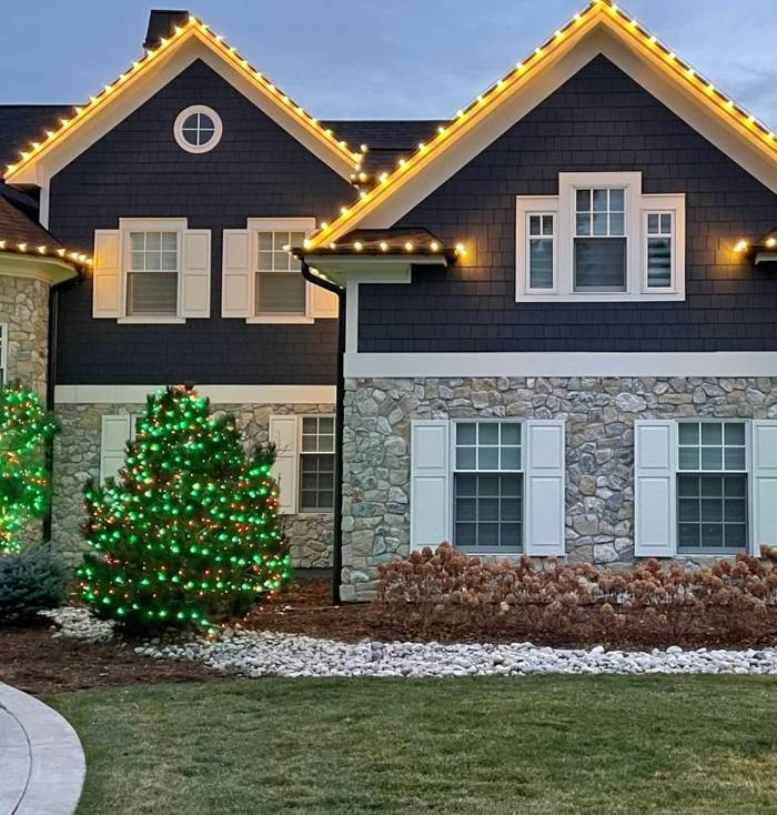 A gabled stone home with warm white roofline lights and a green-lit evergreen at dusk