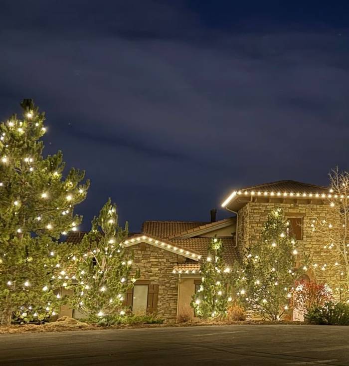Stone home with roofline lights and several lit pines at night