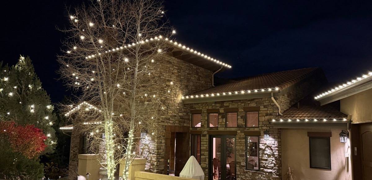 A stone home with warm white roofline lights and wrapped aspens, installed by Sunlight